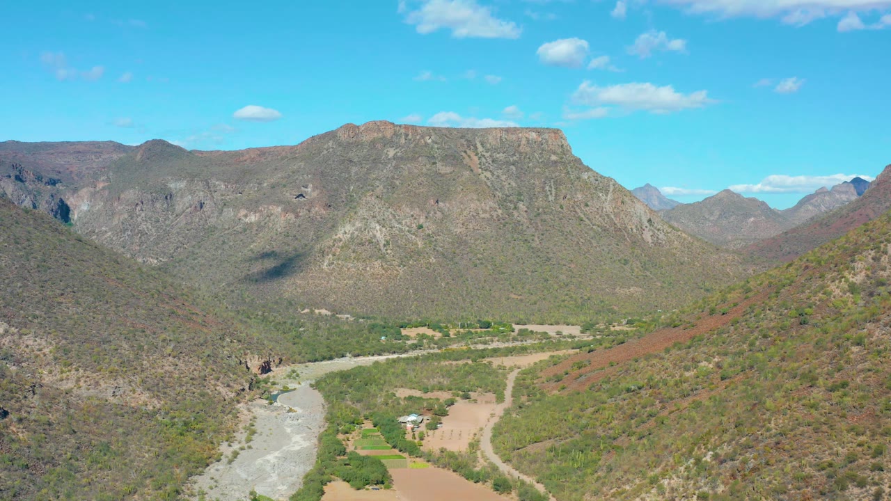 A second aerial view of San Javier, Baja California Sur, with the river channel low in frame, rural roads crossing the valley floor, green patches, dry hillsides, distant mountains, and no visible people.