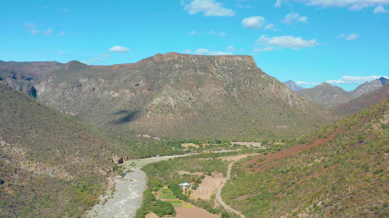 Mid-clip aerial frame in San Javier, Baja California Sur, with a dry foreground hillside, pale riverbed at lower left, small valley structures, green vegetation, a broad rocky ridge, clouds, and no visible people.
