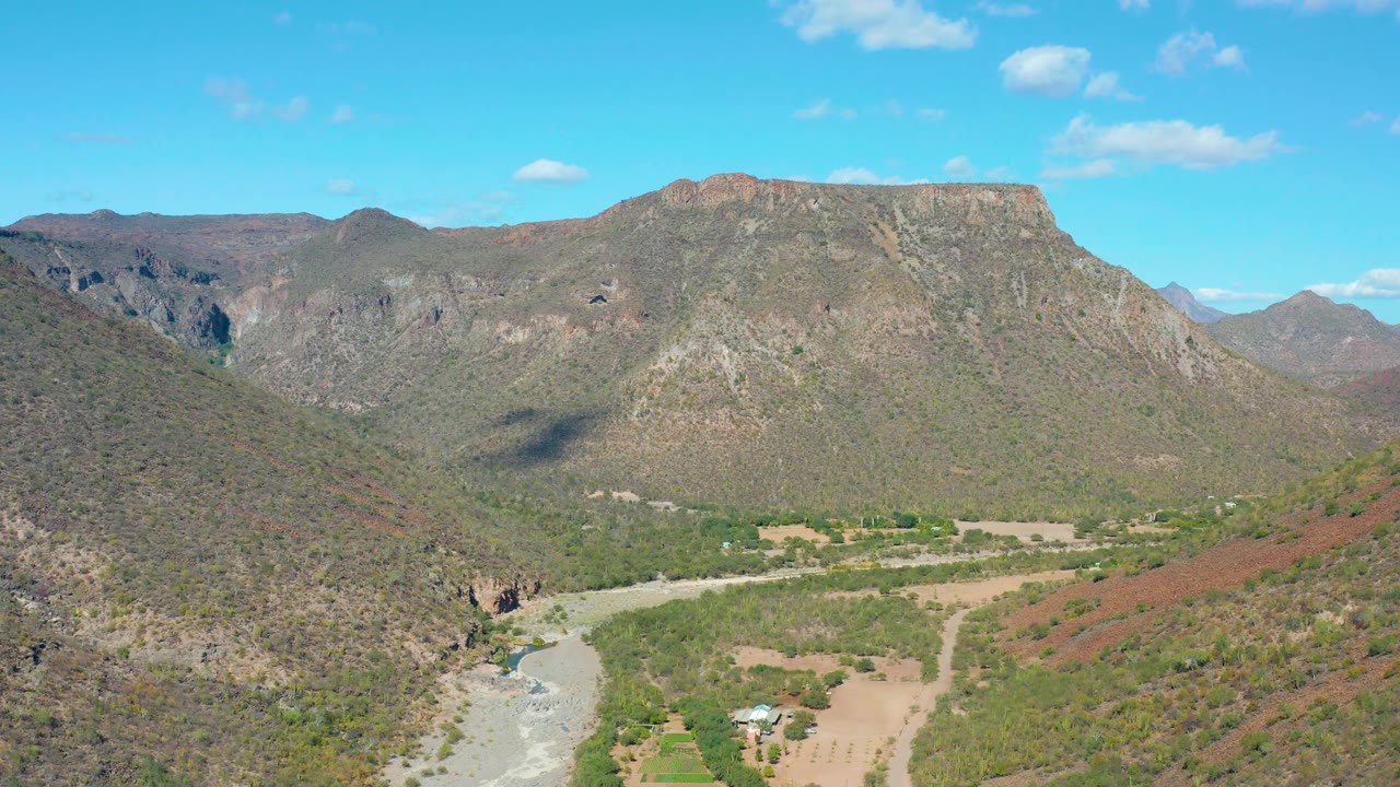 San Javier aerial frame with a broad mountain face ahead, darker shadow on the left slope, riverbed below, scattered greenery, and no visible people.
