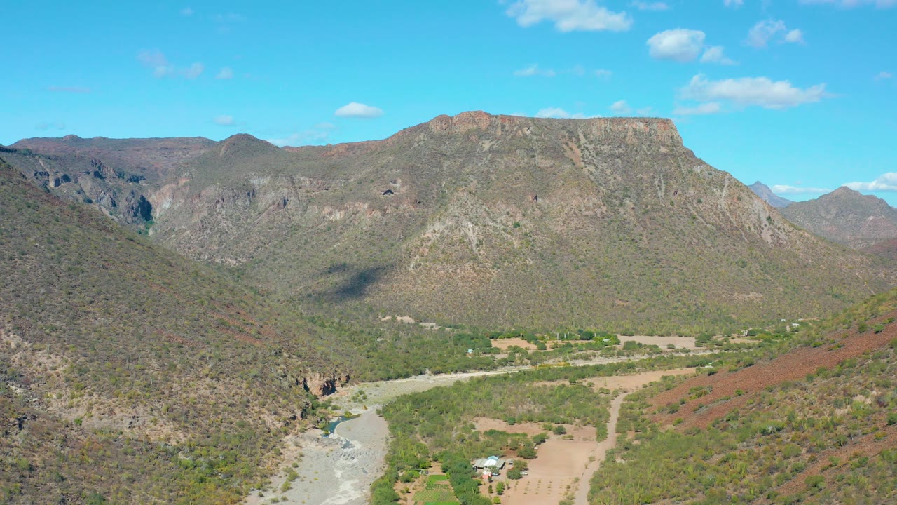 A wide aerial frame over San Javier with the pale riverbed in the lower left, a green and tan valley floor, rocky hillsides, distant ridges, and no visible people.