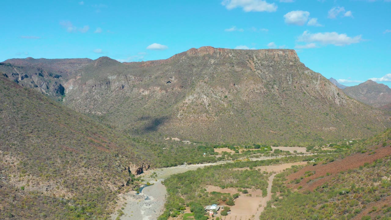 Opening aerial view of San Javier, Baja California Sur with a pale river curving through a green and brown mountain valley under small clouds, no visible people.