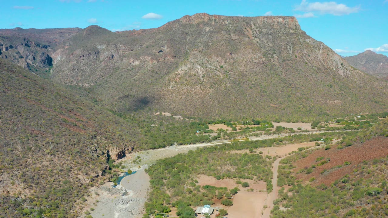 Aerial view over San Javier, Baja California Sur showing a rocky mountain valley, pale riverbed, green vegetation, tan ground, blue sky, and no visible people.