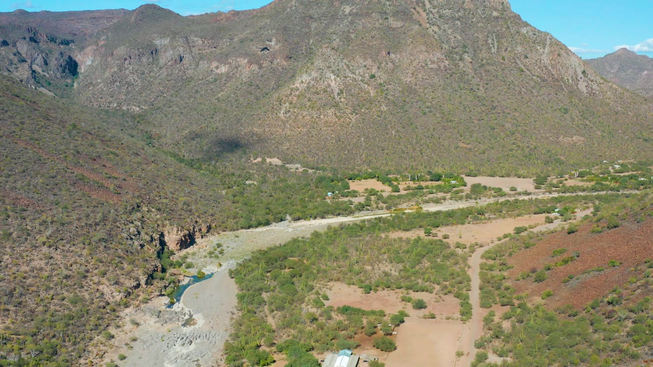 Opening aerial frame over San Javier, Baja California Sur showing rocky mountains, a pale riverbed, scrubby green vegetation, tan dirt clearings, and no visible people.