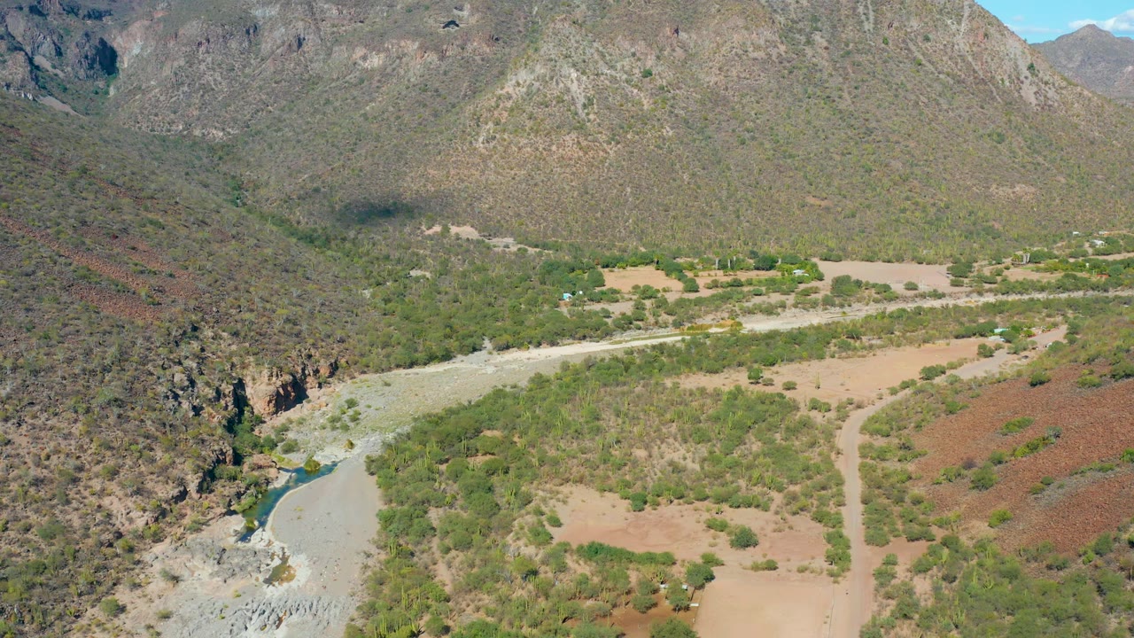 Wide drone view of a dry valley road running near a river and green brush.