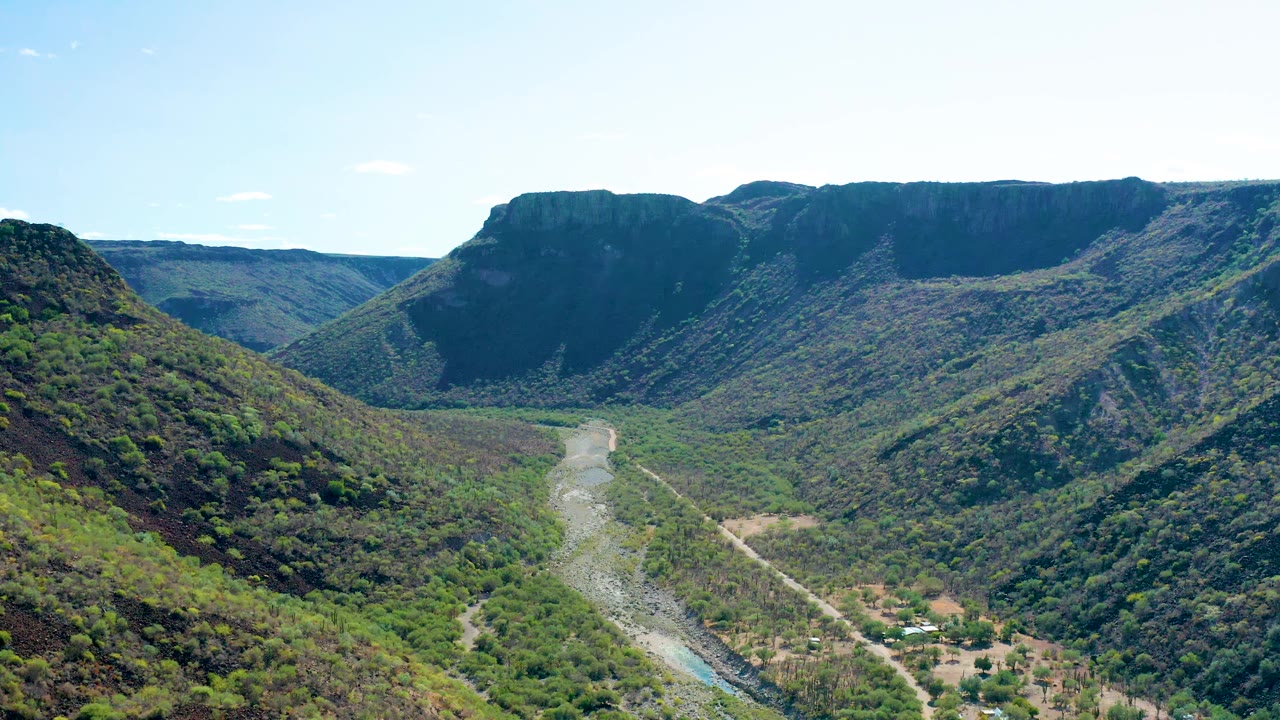 San Javier aerial frame with the riverbed centered low in the valley, a pale road beside it, steep green hillsides, and no visible people.