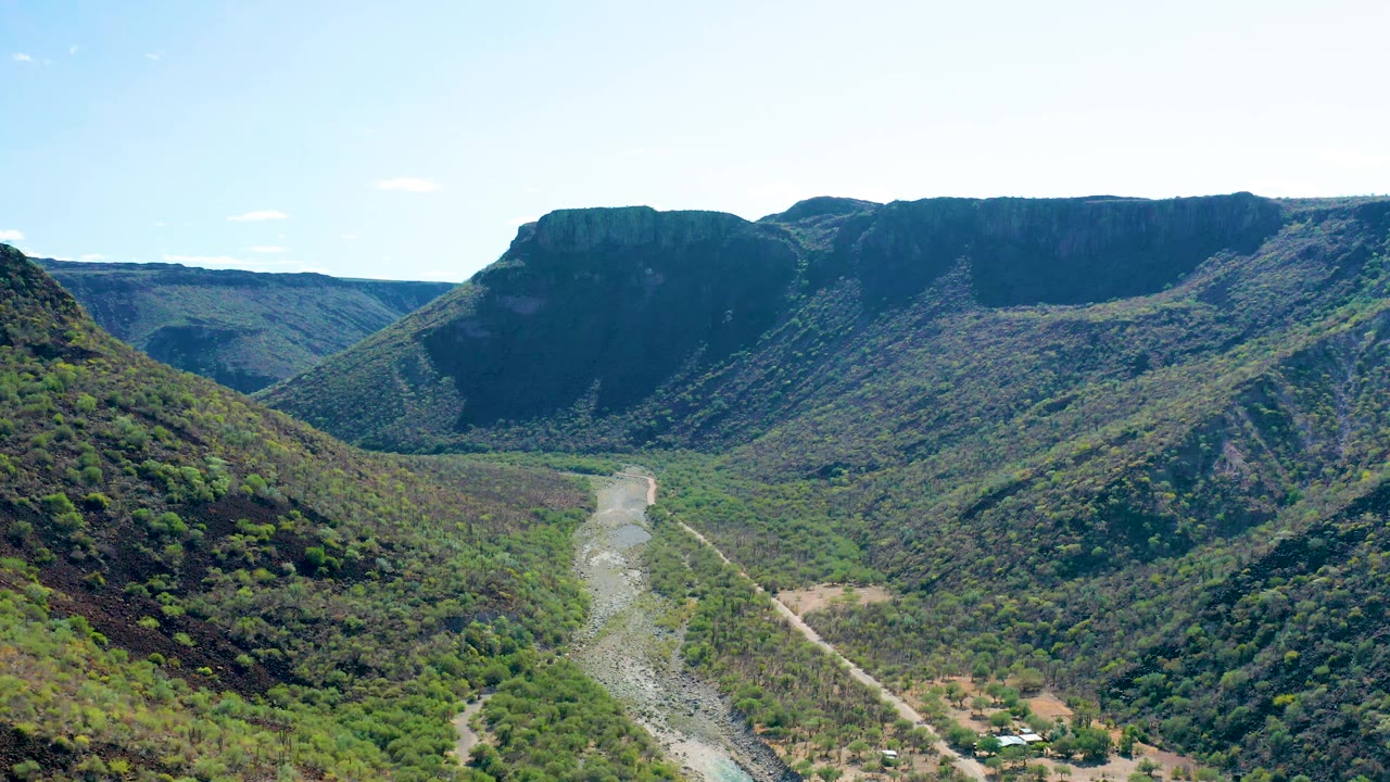 Aerial frame over San Javier, Baja California Sur, with the riverbed centered between green hills, rocky slopes on both sides, bright daylight, and no visible people.