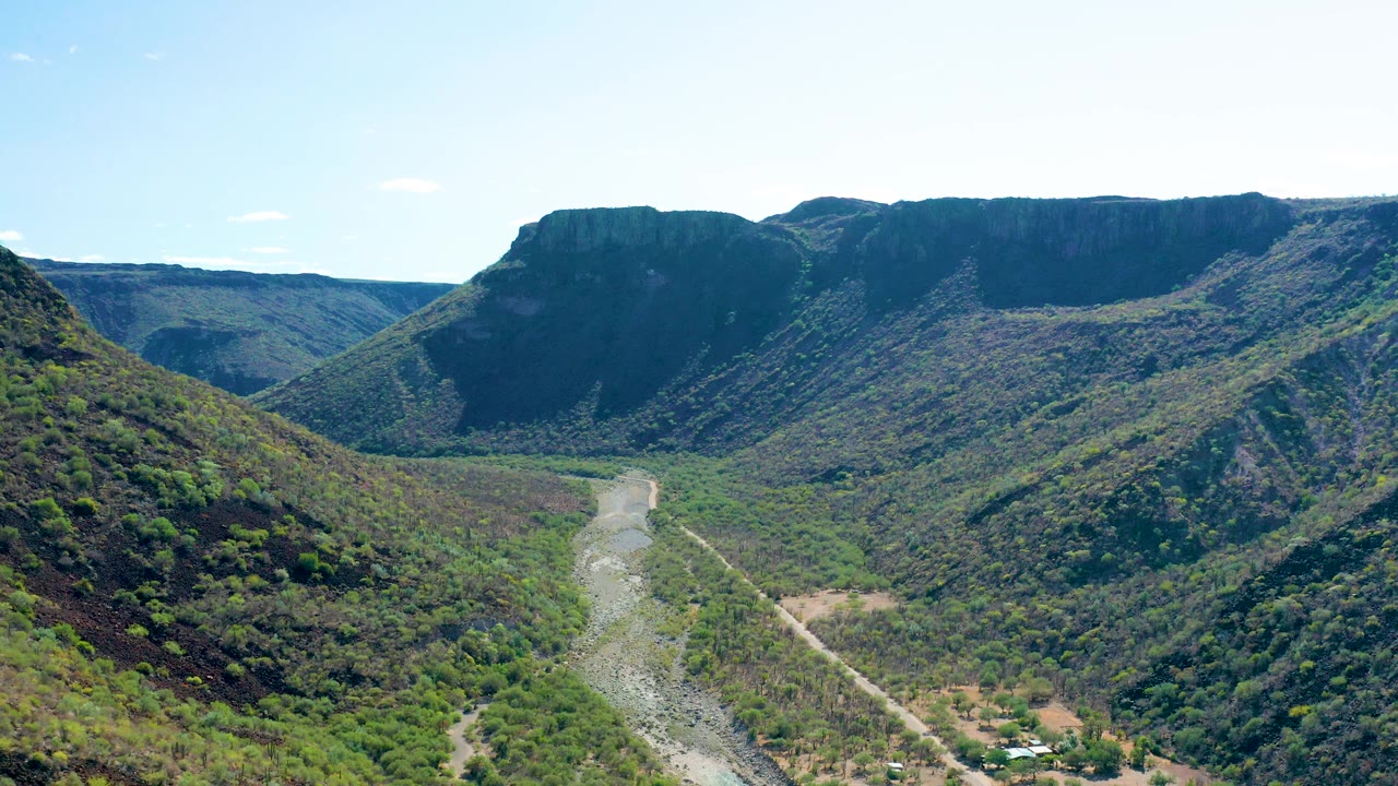 San Javier, Baja California Sur valley seen from above, with a pale riverbed curving through green brush, steep mountain faces in shadow and sun, and no visible people.