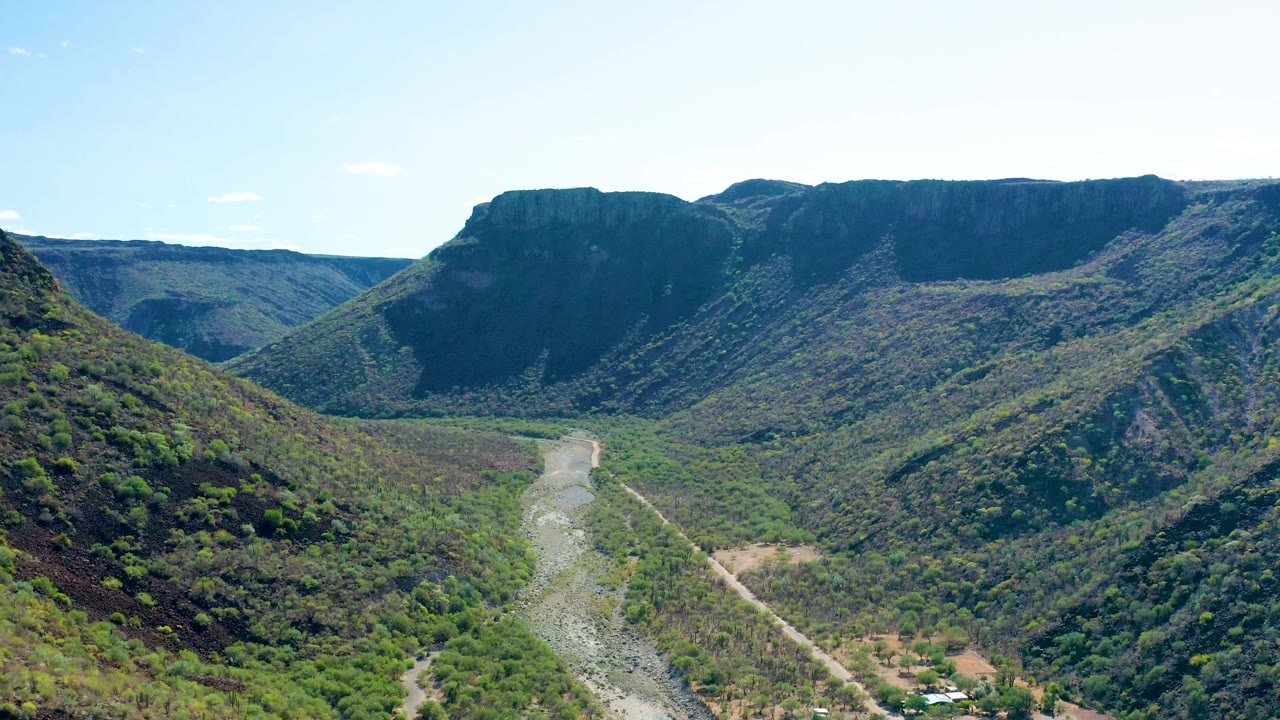 A wide aerial view in San Javier, Baja California Sur, showing the riverbed dropping toward the lower frame, rugged green slopes, distant mesa-like ridges, and no visible people.