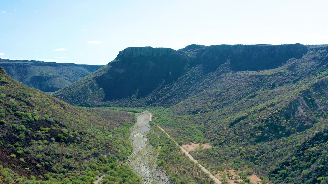 Early frame from San Javier, Baja California Sur, with the river channel centered low in the valley, dense green vegetation on both slopes, and no visible people.
