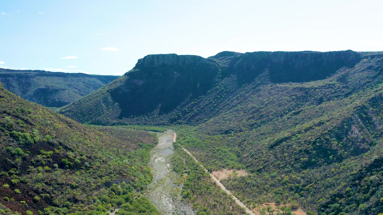 Mid-clip aerial frame over San Javier, Baja California Sur, with a dry-looking pale riverbed, dense shrubs, steep valley walls, and no visible people.