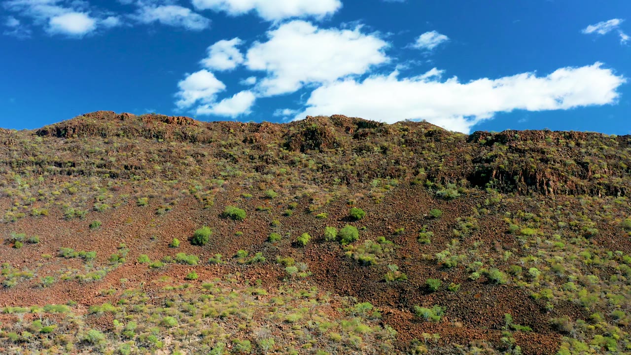 San Javier, Baja California Sur rocky slope with small green shrubs across reddish terrain, a darker ridge section near center, blue sky, clouds, and no visible people.