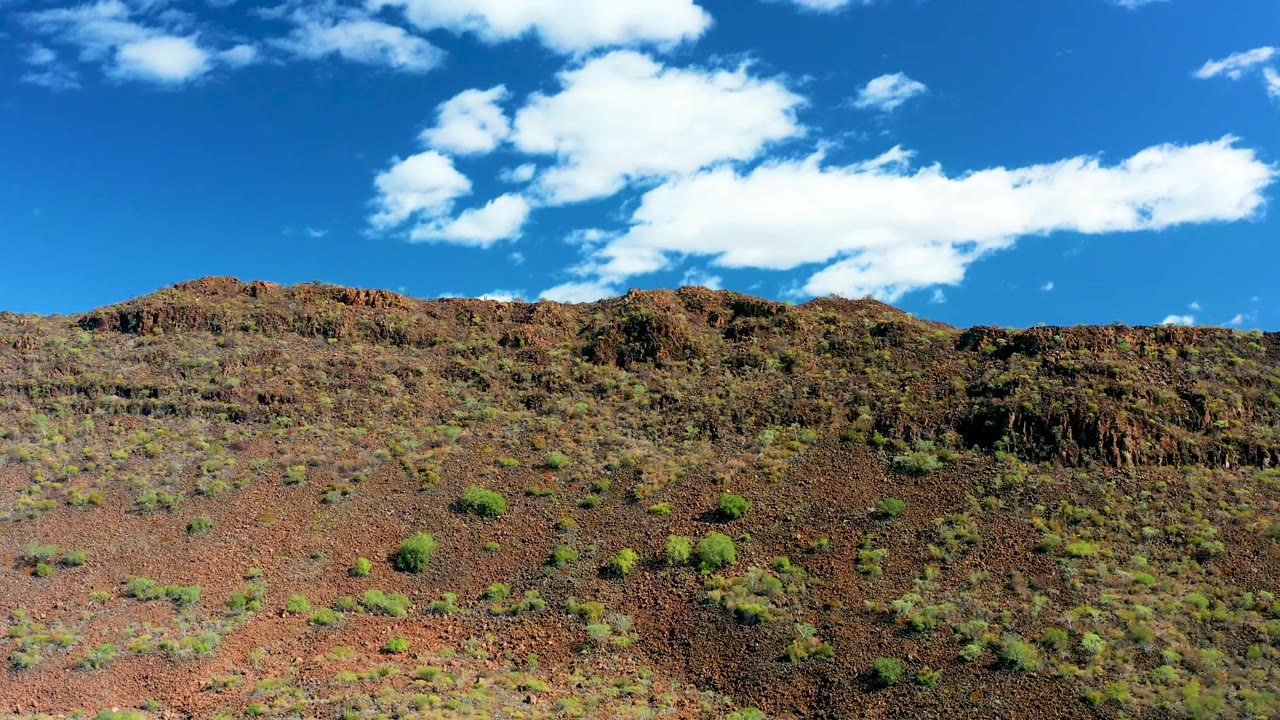 At 0:01 in San Javier, Baja California Sur, the rocky slope fills the lower frame while a large white cloud band stretches across the blue sky, with no visible people.