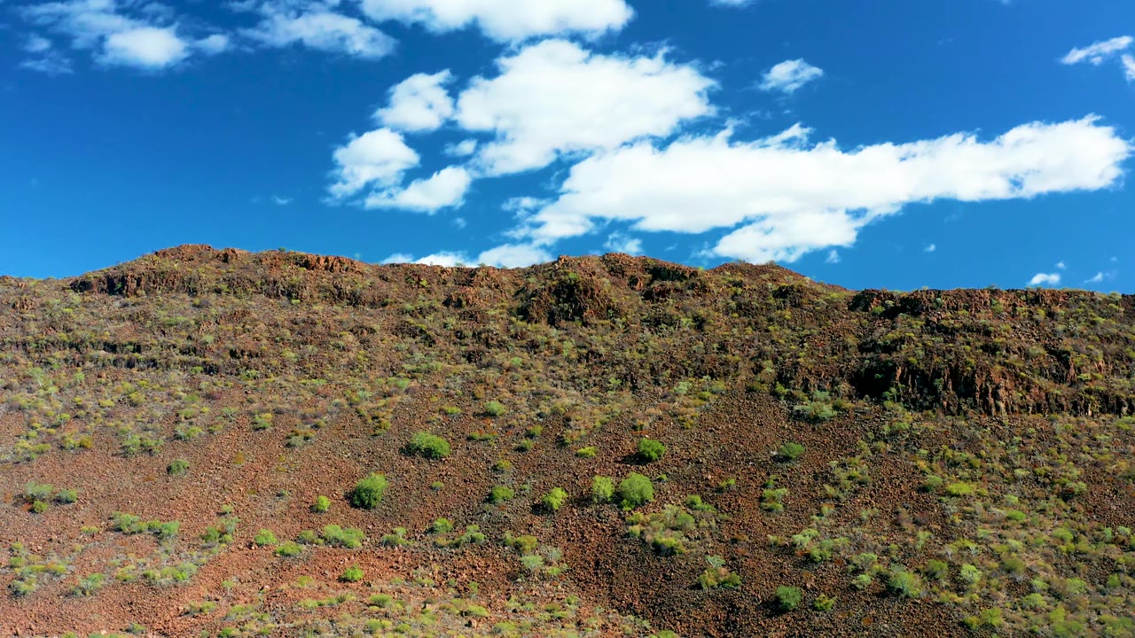 At 0:02 in San Javier, Baja California Sur, the ridge line runs across the frame above patchy green shrubs on reddish rock, with bright clouds against blue sky and no visible people.