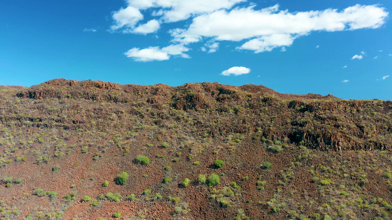 San Javier desert ridge with reddish rock, small green shrubs across the slope, bright white clouds overhead, and no visible people one second into the clip.