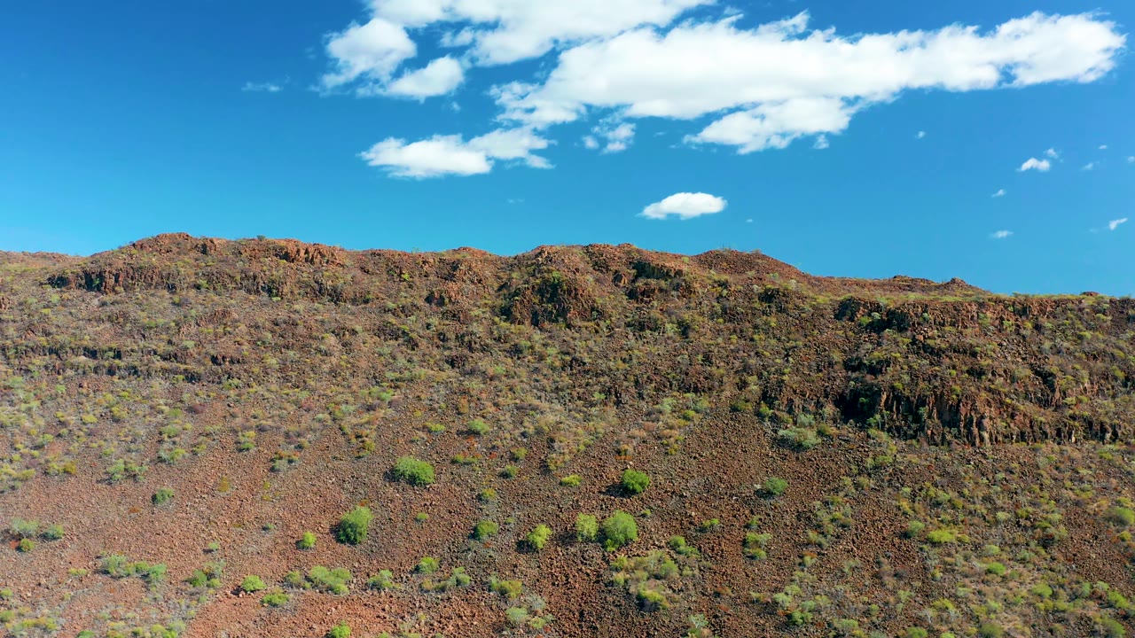 San Javier, Baja California Sur arid slope with rough brown rock, scattered green bushes, a high blue sky, drifting white cloud shapes, and no visible people.