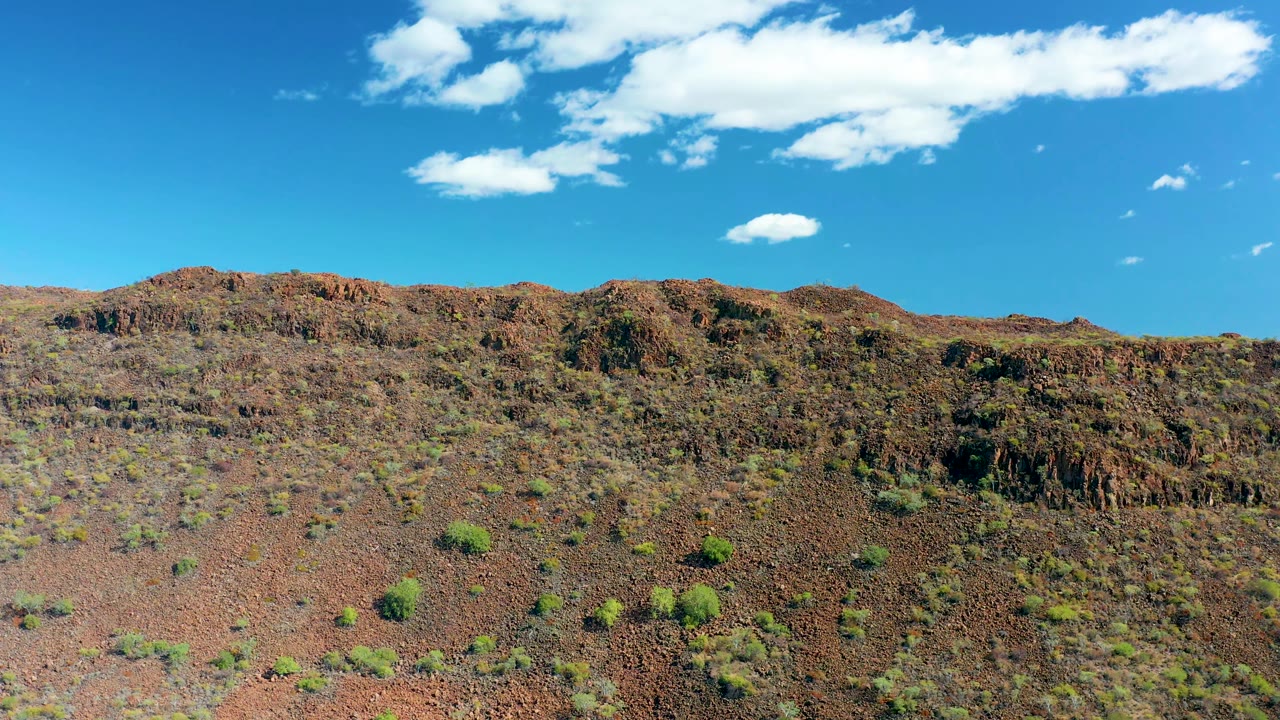 San Javier desert hill with rocky ledges, sparse green vegetation on the lower slope, white clouds near the top of the frame, and no visible people.