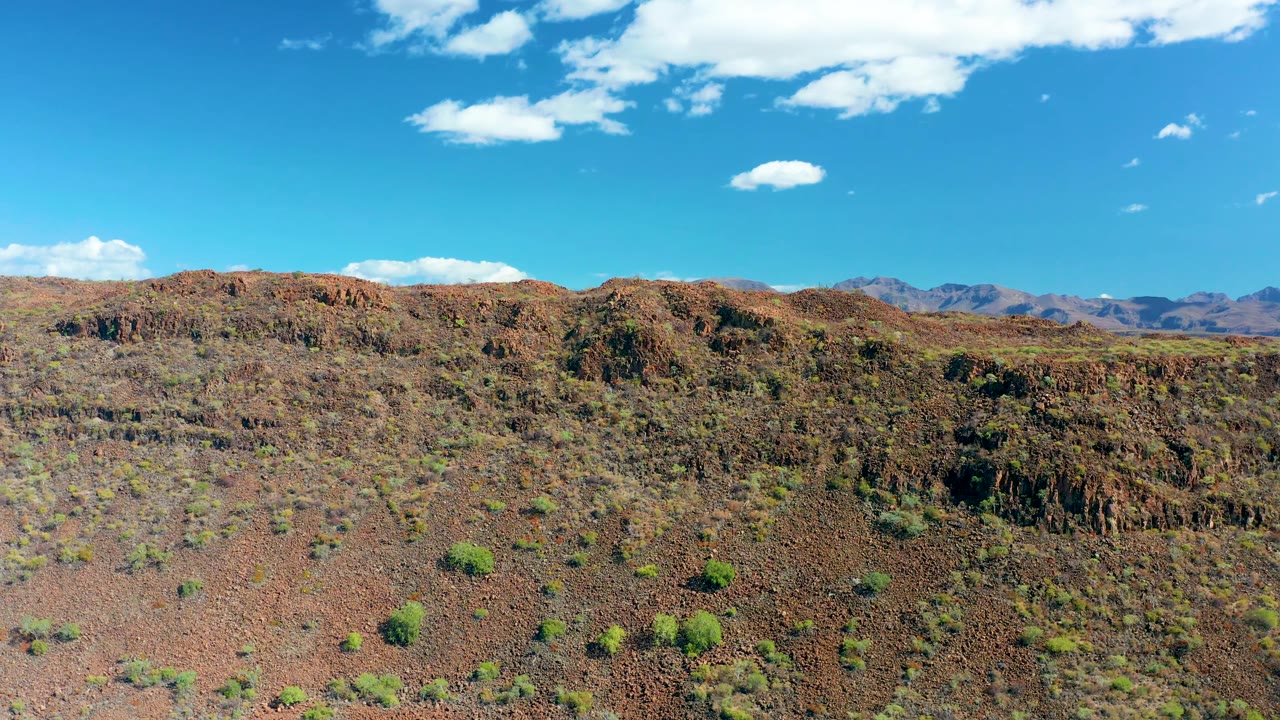 Aerial view over red-brown rocky desert near San Javier, Baja California Sur, with scattered green shrubs, distant mountains, white clouds, and no visible people.