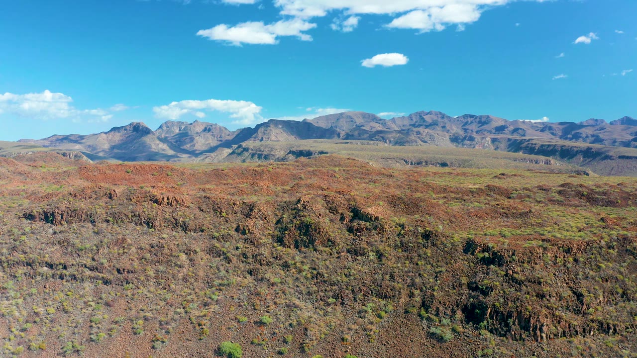 Early frame in San Javier, Baja California Sur with a broad rocky plateau, reddish scrub-covered ground, a layered mountain range, bright blue sky, and no visible people.