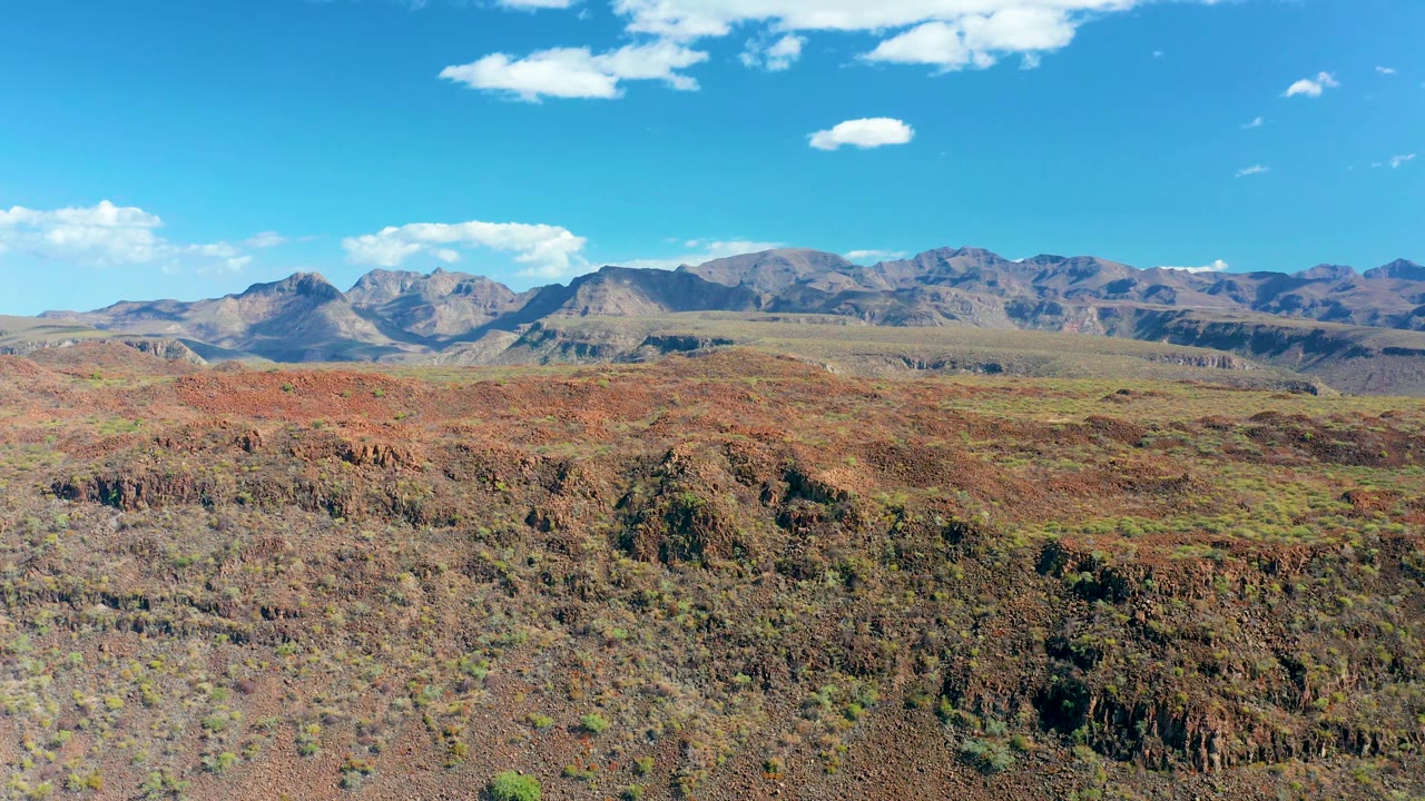 San Javier, Baja California Sur frame with rough red-brown desert ground filling the lower half, a dry valley beyond it, distant ridgelines, small clouds, and no visible people.