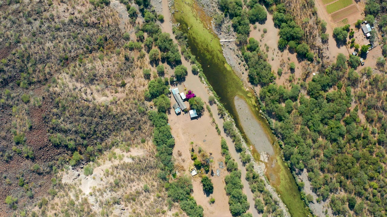 Overhead aerial frame in San Javier, Baja California Sur showing a green irrigation canal, dry scrubland, dirt tracks, small buildings, and no visible people.