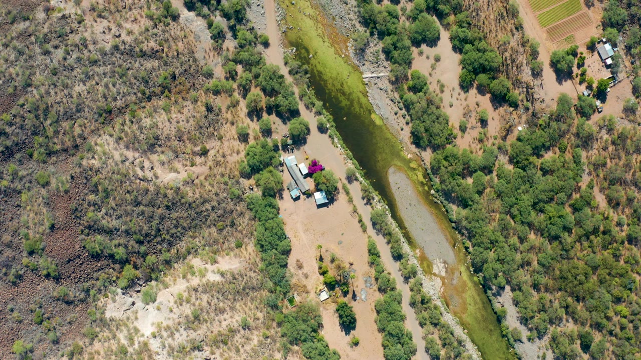 High overhead frame in San Javier, Baja California Sur showing dry hillside texture, green canal water, tree clusters, rectangular field plots, small buildings, and no visible people.