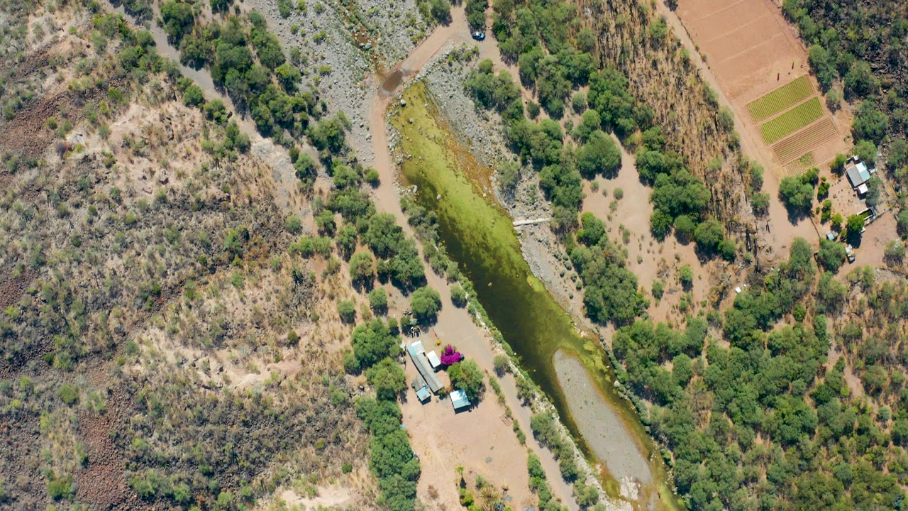 Overhead frame from San Javier, Baja California Sur showing a narrow green canal, dry brush, scattered trees, dirt tracks, small structures, and no visible people.