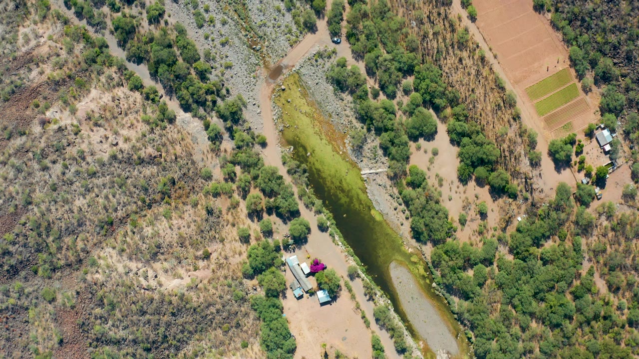 Drone frame from San Javier, Baja California Sur showing the green canal near a dirt road, rectangular field plots, small rural structures, parked vehicles, and no visible people.