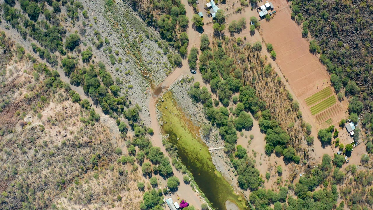 Second aerial frame over San Javier shows the same river bend with green floodwater, exposed pale riverbed, brown cultivated plots, tree clusters, buildings, and no visible people.
