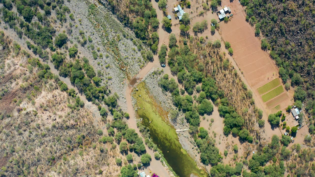 Aerial frame in San Javier with greenish floodwater running beside pale stone banks, dry scrub slopes, rectangular brown fields, trees, buildings, and no visible people.