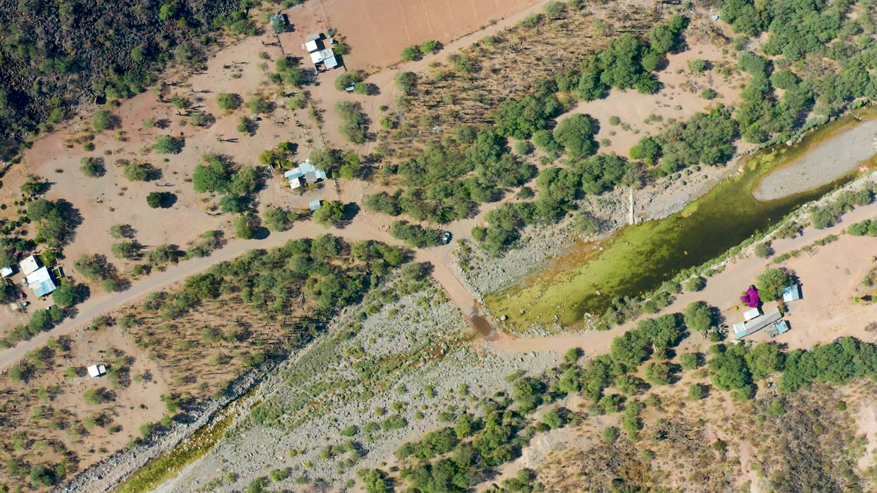 High overhead aerial frame in San Javier, Baja California Sur showing a greenish canal bordered by dry tan ground, scrub vegetation, dirt tracks, scattered small buildings, and no visible people.