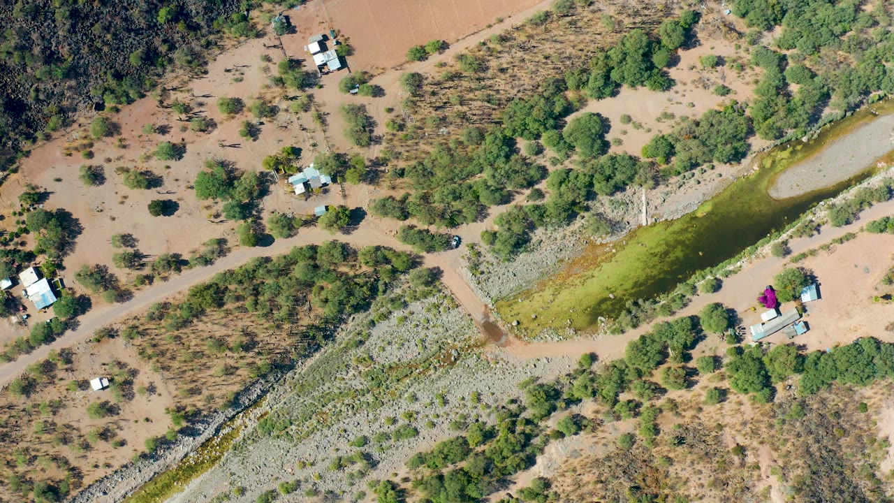 A near-vertical aerial view in San Javier shows the narrow green canal beside exposed earth, low scrub, tree cover, dirt paths, several small structures, and no visible people.