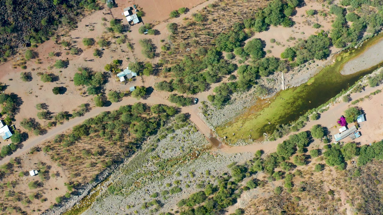 Drone view of a dry campsite area with dirt tracks, trees, a green strip, and small structures.
