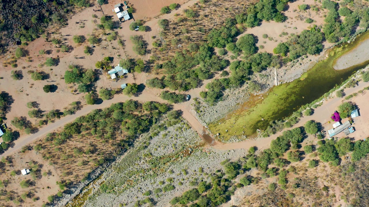 Aerial view in San Javier, Baja California Sur with the dirt road and green canal crossing diagonally, the white vehicle slightly farther along the road, and no visible people.