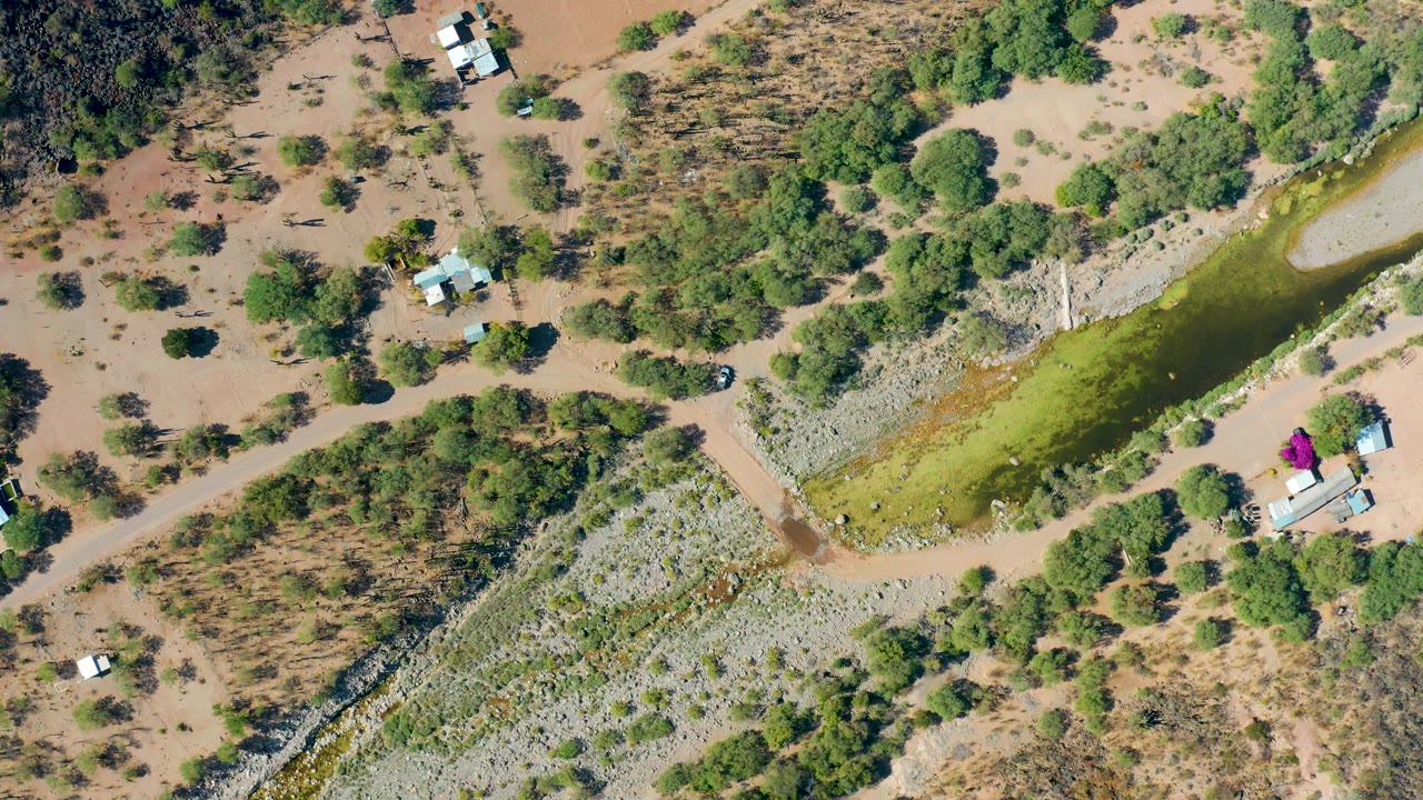 Top-down San Javier aerial showing the green canal bordered by dry earth, a diagonal dirt road, scattered trees, light-roofed structures, and no visible people.