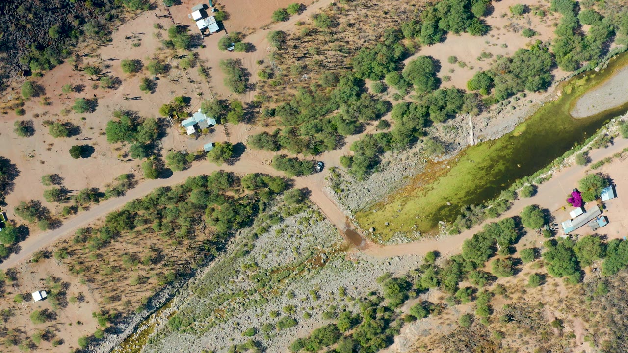 Drone frame of a rural dirt road near a green canal and small structures in dry brush.
