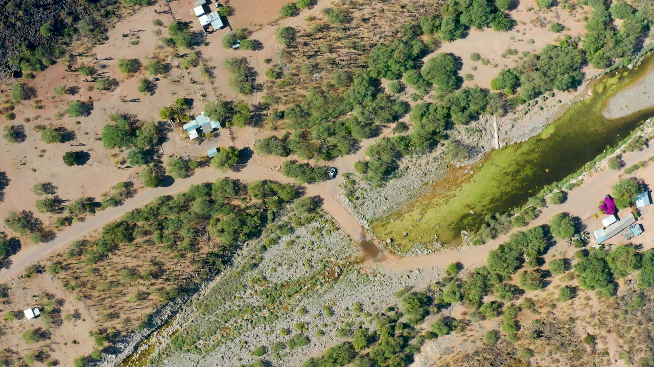 Opening overhead frame in San Javier, Baja California Sur, showing a narrow green canal, dirt tracks, scattered trees, small structures, parked vehicles, and no visible people.