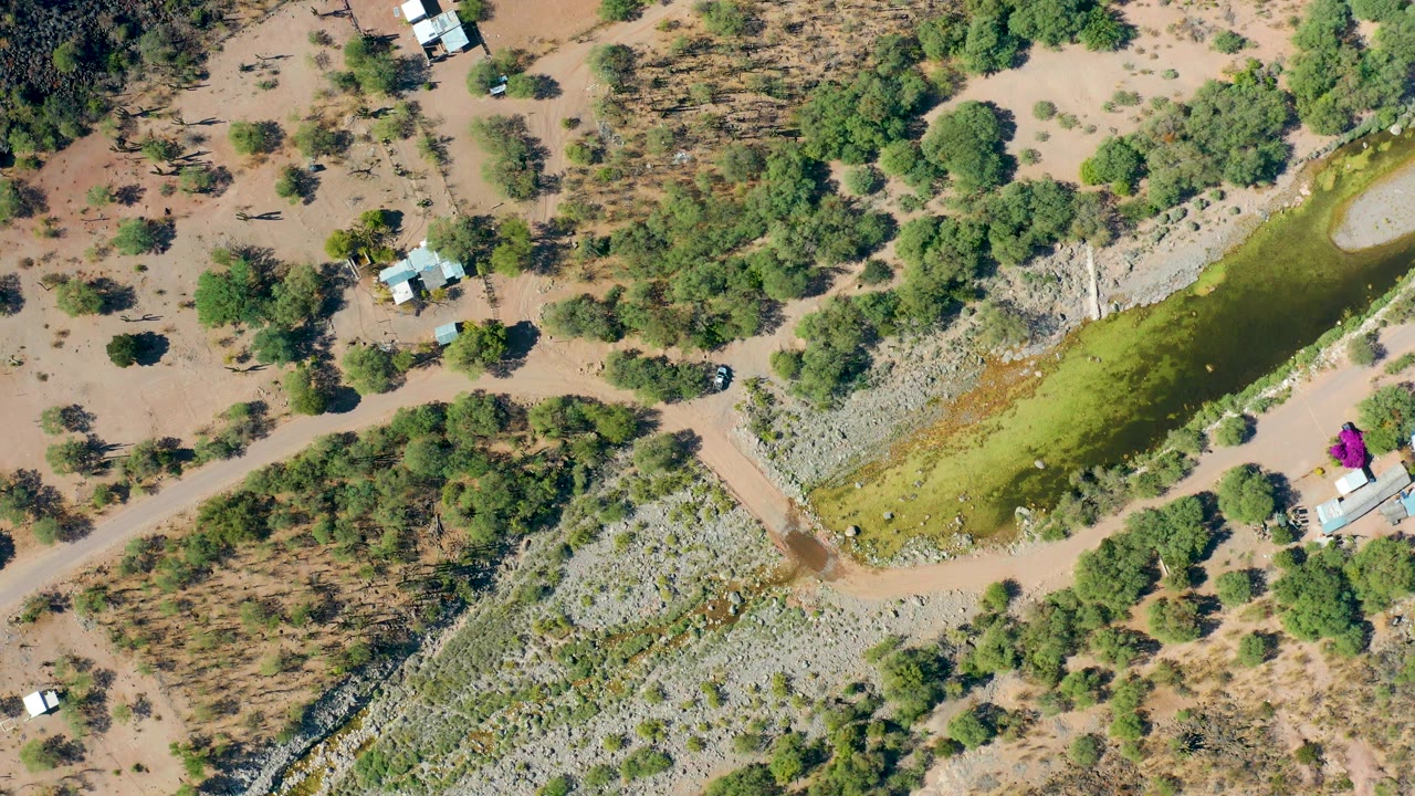 Aerial frame in San Javier, Baja California Sur with the green water body running diagonally, dry brushland around it, small structures nearby, and no visible people.