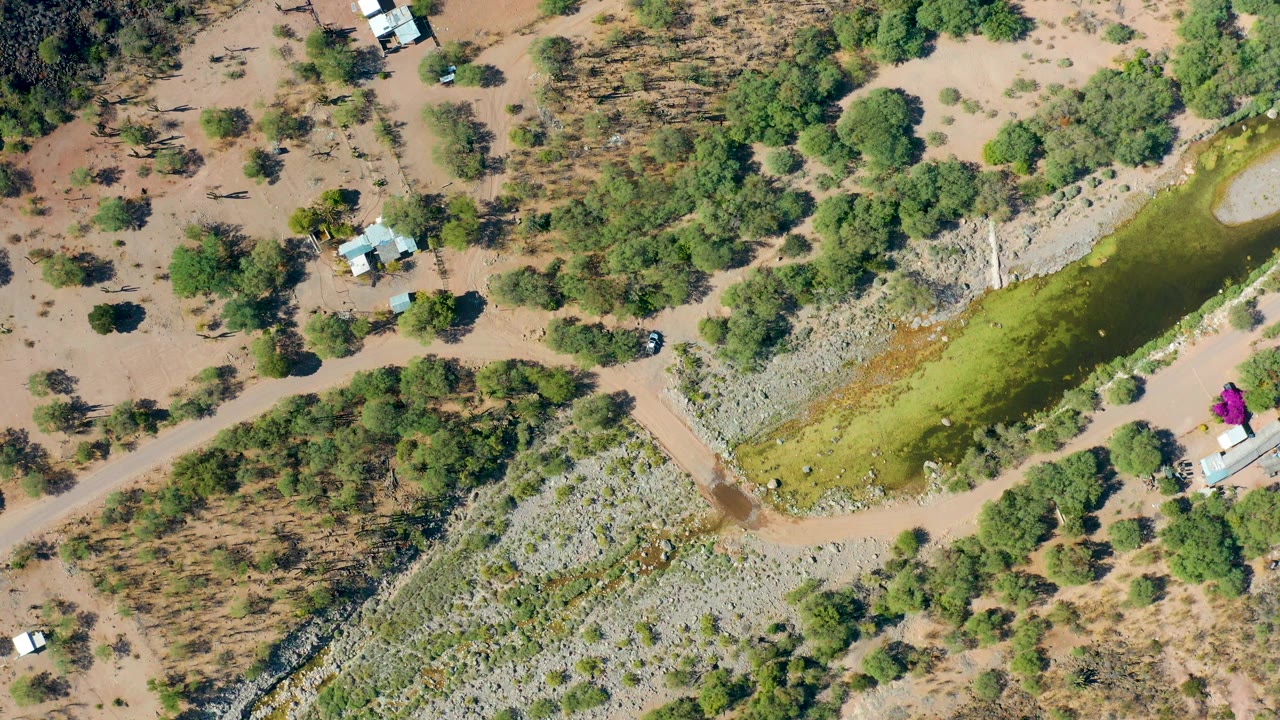 Overhead rural scene with green water, dirt road, dry vegetation, and small vehicles.