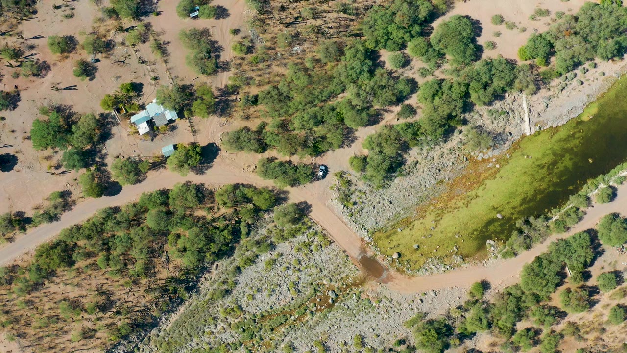 Overhead frame from San Javier, Baja California Sur showing green-brown floodwater beside pale rocks, dirt roads, scattered trees, a small house, and no visible people.