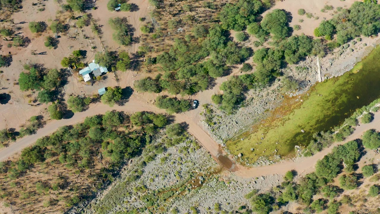 Top-down view of floodwater bordered by rocks, trees, dirt tracks, and a small house.