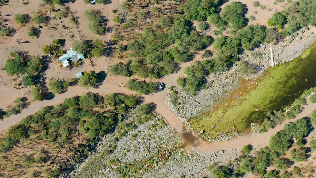 Overhead frame from San Javier, Baja California Sur showing a dirt road beside green water, rocky banks, scattered trees, small rural structures, one dark vehicle, and no visible people.