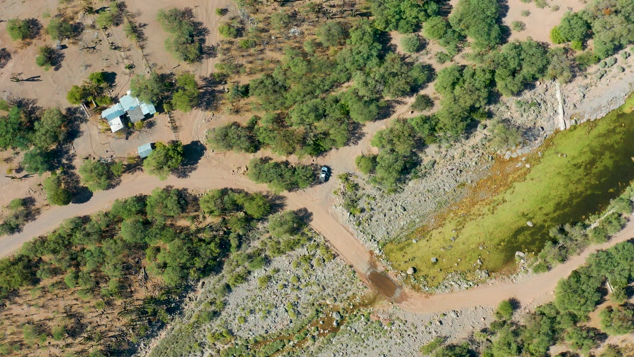 Aerial frame of muddy water, diagonal dirt road, rocky bank, trees, small building, and vehicle.