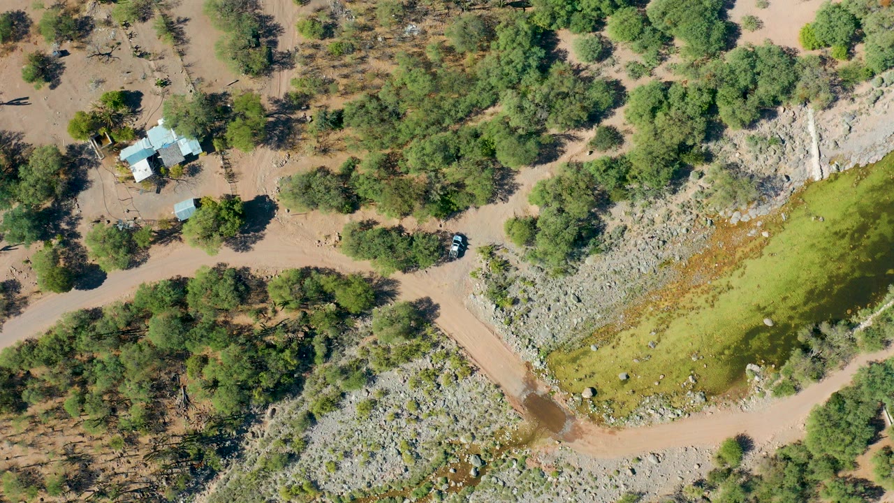 Overhead frame in San Javier, Baja California Sur showing a dirt road beside a small house compound, rocky ground, trees, green floodwater, and no visible people.
