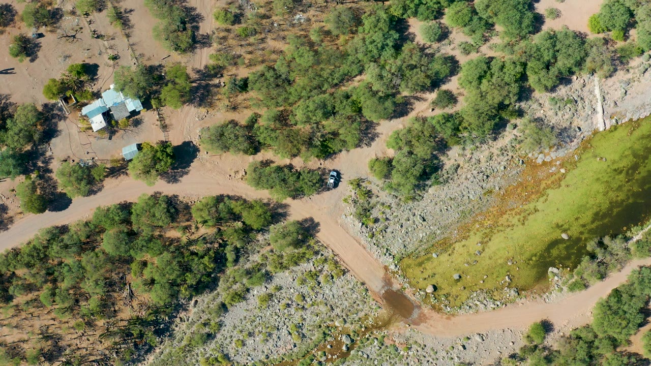 Overhead rural flood scene with a dirt road crossing muddy water, trees, rocks, and a house.