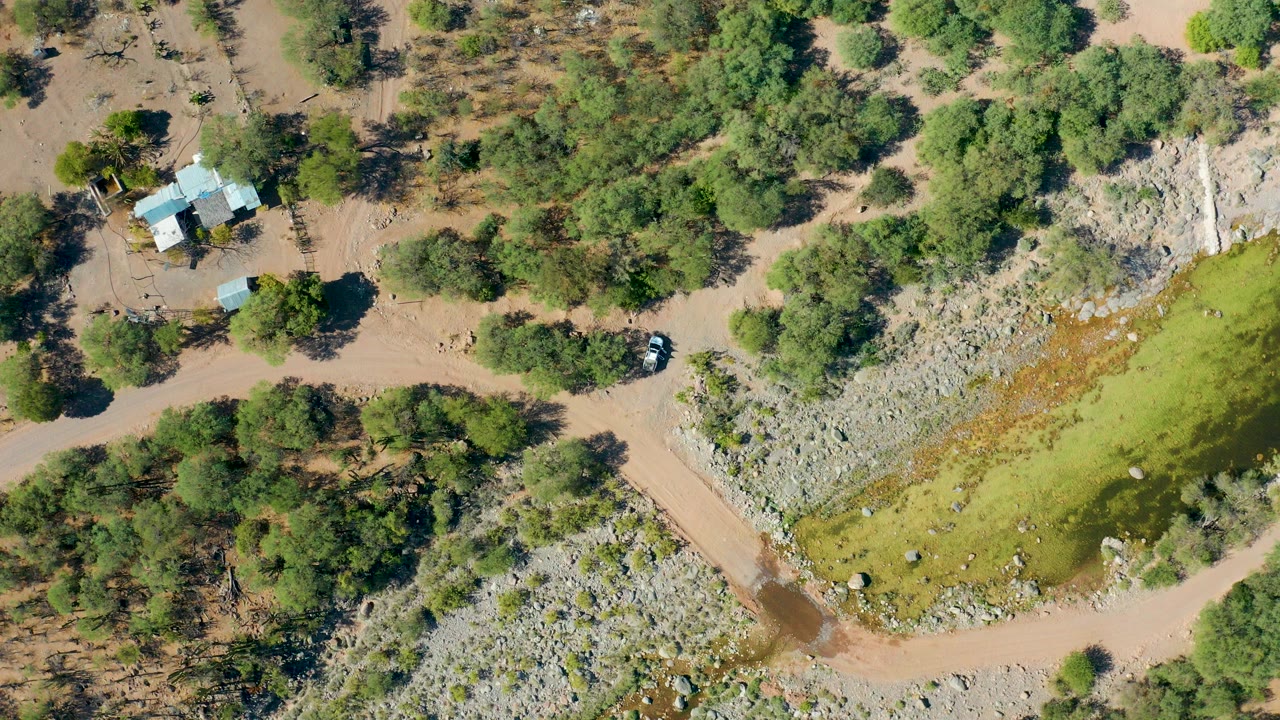 Overhead view in San Javier, Baja California Sur, showing small buildings, a parked car, dirt roads, rocky terrain, trees, and green-brown floodwater with no visible people.