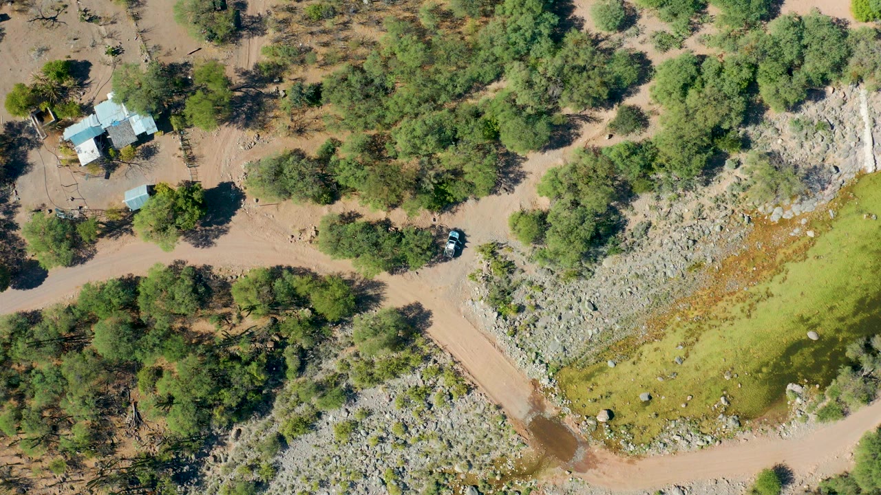 Overhead drone frame in San Javier, Baja California Sur showing a dirt road beside a small light-roofed residence, dense green trees, rocky ground, and green-brown floodwater at the lower right with no visible people.