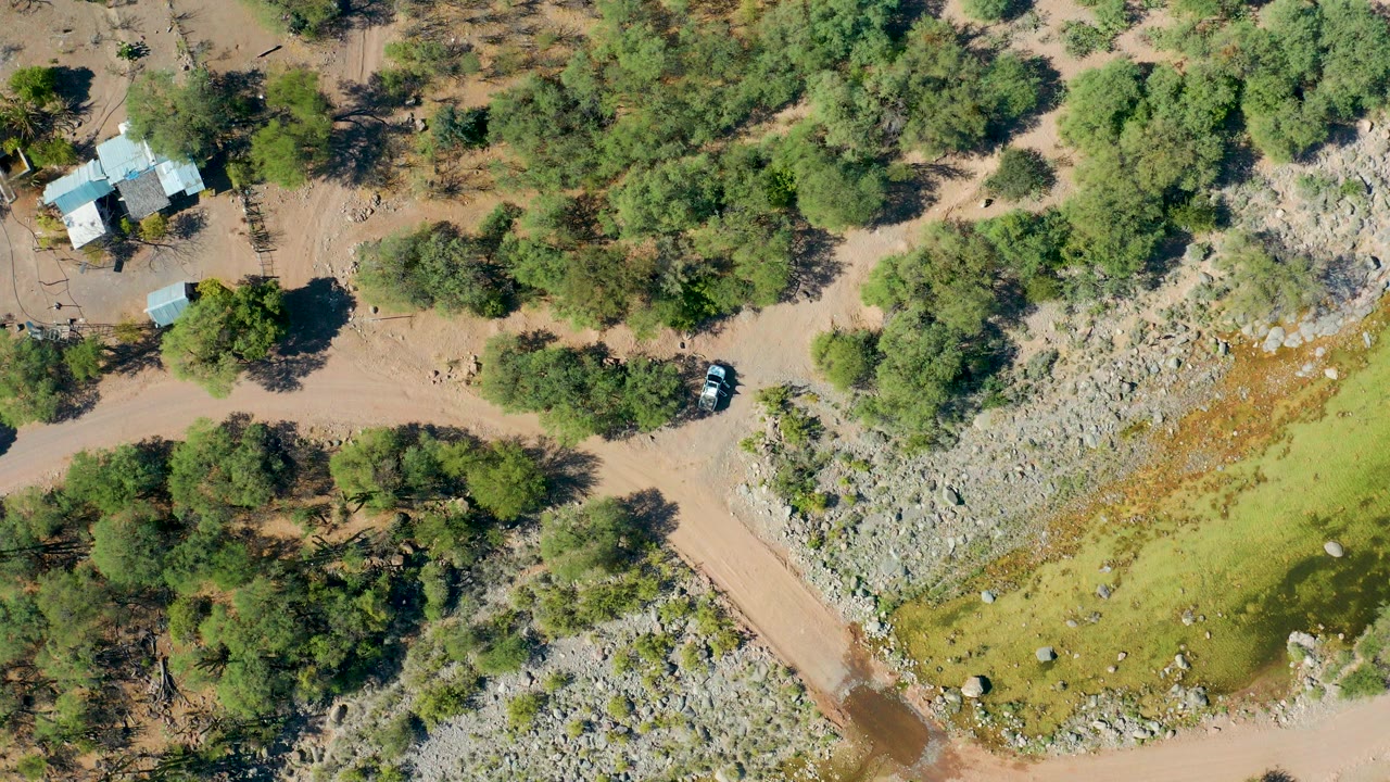 Overhead view in San Javier, Baja California Sur, showing a dirt road crossing a dry riverbed, scattered green trees, a small light-roofed building, a vehicle, and no visible people.