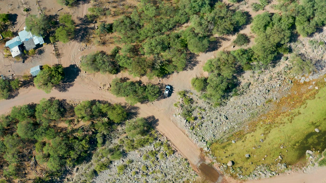 Overhead landscape with a dirt crossing, green trees, a dry wash, and a small building.