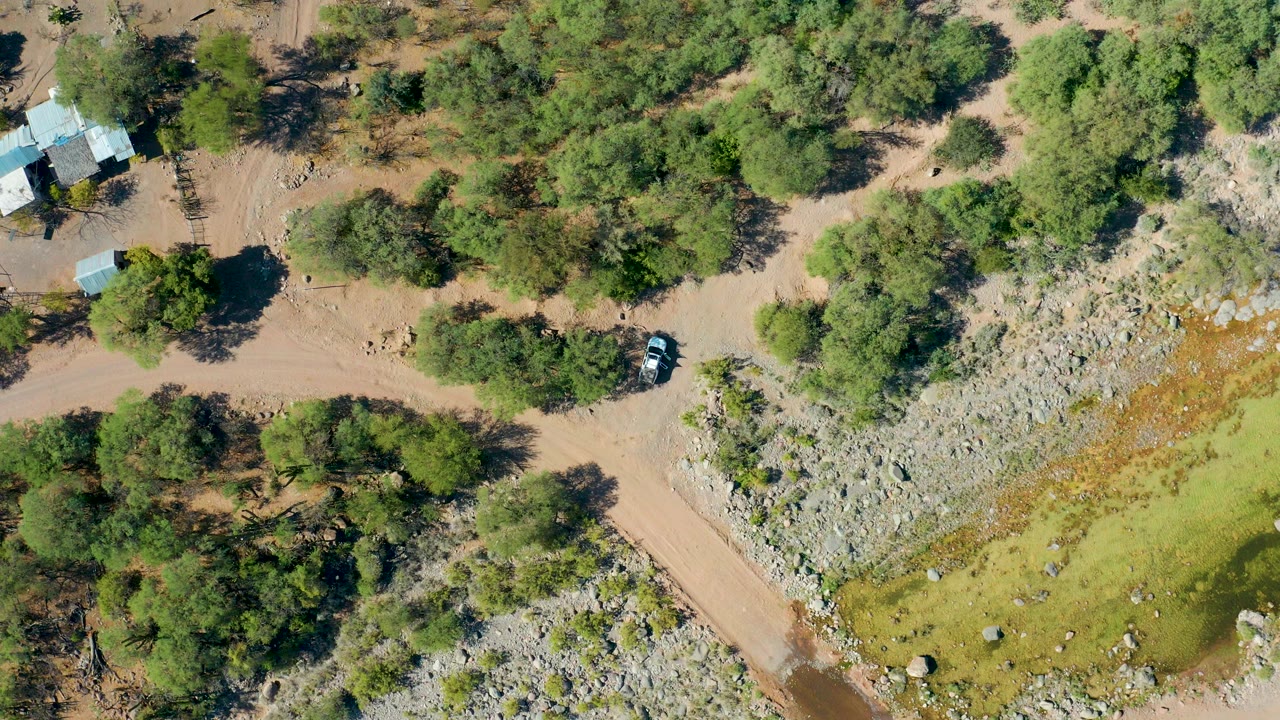Overhead view in San Javier, Baja California Sur of a tan dirt road curving past trees, rocky open ground, a small roofed structure, and a parked vehicle, with no visible people.