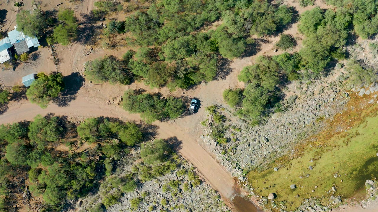 Aerial frame over San Javier, Baja California Sur showing the dirt road running between dense green trees and rocky pale ground, with a small structure near the upper edge and no visible people.